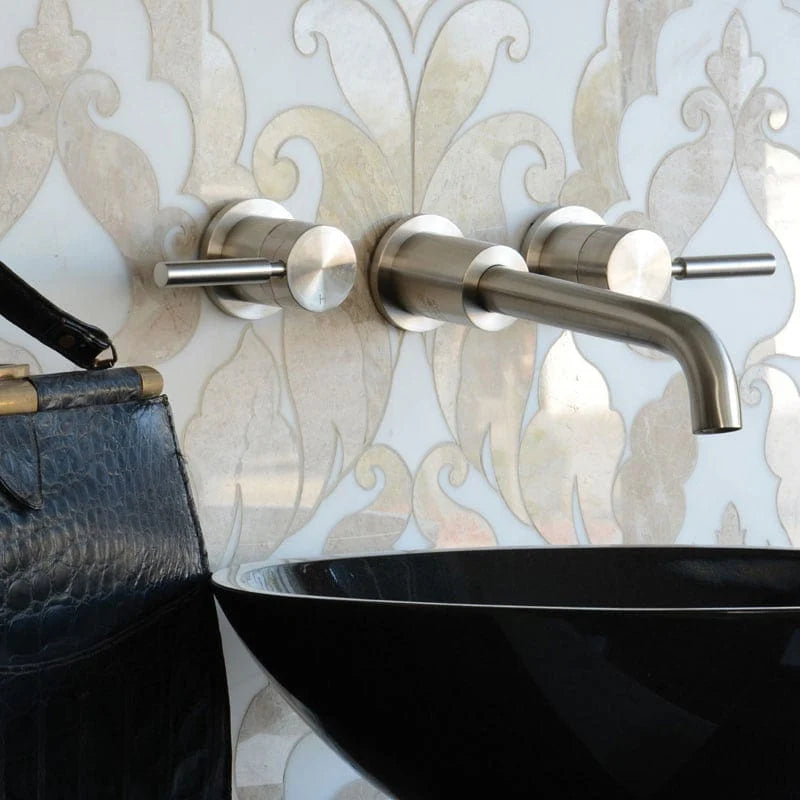 A modern wall-mounted bathroom faucet set with two cylindrical handles and a curved spout in brushed silver metal, installed on a white damask-patterned wall above a black vessel sink. Part of a dark textured bag is visible on the left.