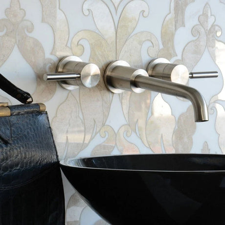 A modern wall-mounted bathroom faucet set with two cylindrical handles and a curved spout in brushed silver metal, installed on a white damask-patterned wall above a black vessel sink. Part of a dark textured bag is visible on the left.
